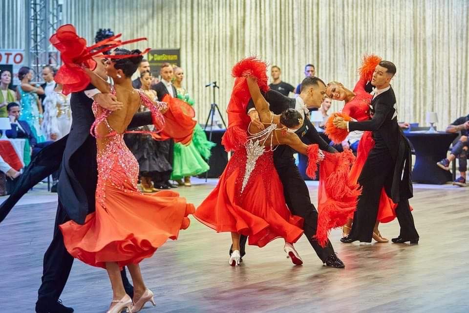 A Standard lady’s dress adds greatly to the couple’s overall impression. Here, WDSF World Adult 10-Dance Vice Champions Earle Williamson and Veronika Myshko (center) dance alongside two other couples at the 2022 World Championship event in Bratislava, Slovakia. Although all three pictured dresses are red, they are each notably unique. Photo Courtesy of Earle Williamson and Veronika Myshko.